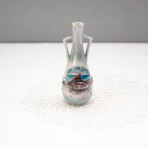 May include: A small, pearlescent ceramic vase with two handles. The vase features a colorful image of the Sea Lion Caves, with text. The rim is trimmed with gold. The vase is displayed on a white, lacy doily.
