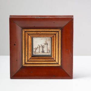 May include: A square wooden picture frame with a deep reddish-brown outer border and a lighter, multi-layered inner border in natural wood tones. The frame contains a small, centered black and white photograph depicting a historic stone castle.