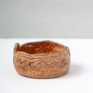 May include: A small, round, ceramic bowl with a textured, wood-like exterior. The bowl is primarily brown with orange accents inside. The rim has a slight lip. The bowl is likely handmade and suitable for small items or as a decorative piece.