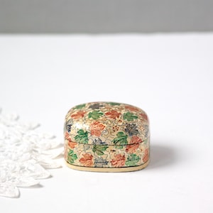 May include: A small, oval-shaped decorative box with a lid. The box is covered in a pattern of colorful leaves in shades of red, green, and orange on a cream-colored background. The box has a light beige base.