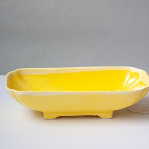 May include: A rectangular, yellow ceramic bowl with a glossy finish. The bowl has a slightly curved shape and sits on four small feet. The interior is bright yellow, and the rim has a cream-colored trim. The bowl is likely used for serving food or as a decorative piece.