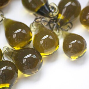 K&ouml;nnte beinhalten: Eine Ansammlung von olivgr&uuml;nen, tropfenf&ouml;rmigen Glasanh&auml;ngern mit kleinen Schlaufen zum Aufh&auml;ngen. Die Anh&auml;nger haben eine transparente, gl&auml;nzende Oberfl&auml;che und sind auf einer wei&szlig;en Oberfl&auml;che verstreut. Das Glas hat einen leichten Gelbstich am Boden.