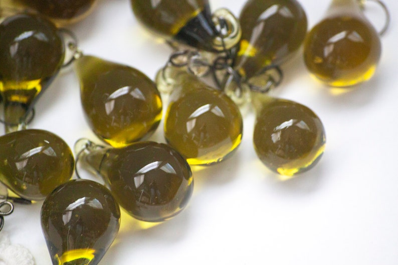 K&ouml;nnte beinhalten: Eine Ansammlung von olivgr&uuml;nen, tropfenf&ouml;rmigen Glasanh&auml;ngern mit kleinen Schlaufen zum Aufh&auml;ngen. Die Anh&auml;nger haben eine transparente, gl&auml;nzende Oberfl&auml;che und sind auf einer wei&szlig;en Oberfl&auml;che verstreut. Das Glas hat einen leichten Gelbstich am Boden.