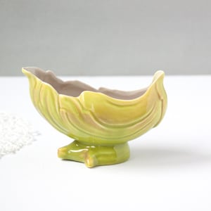 May include: A ceramic, leaf-shaped bowl in shades of green and yellow, with a light brown interior. The bowl sits on a pedestal base with a star-like design. The bowl has a glossy finish and is likely vintage.