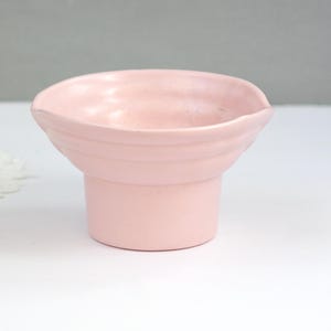 May include: A light pink ceramic pedestal bowl with a rounded top and a cylindrical base. The bowl has a smooth, matte finish and a slightly textured rim. The bowl is designed for decorative use.