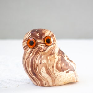 May include: A ceramic owl figurine with a marbled brown and cream pattern. The owl has large, round orange eyes and a slightly tilted head. The figurine is a decorative item.