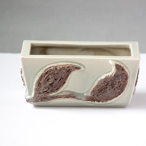 May include: A rectangular, light green ceramic planter with a brown, textured leaf design on the front. The planter has a square shape and a hollow interior. The background is a light gray.