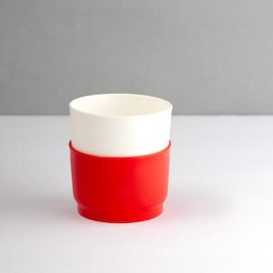 May include: A white ceramic cup with a red silicone sleeve. The cup is cylindrical with a smooth surface. The red sleeve wraps around the bottom half of the cup, providing a contrasting color and a secure grip.