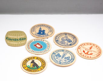 May include: A collection of vintage beer coasters and a gin label. The coasters feature various brewery logos and text, including "Klosterbrauerei Koblenz" and "Adler Bier Altenstadt". The gin label reads "Curtis Gin" and "Smoother Because".