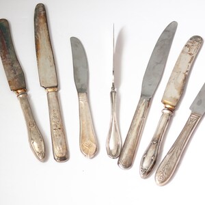May include: A collection of vintage butter knives with silver blades and ornate handles. The knives show signs of age and wear, with some blades displaying discoloration. The handles feature various decorative patterns and designs.