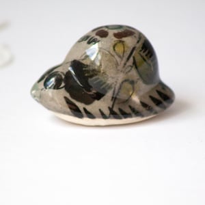 May include: A small, ceramic sculpture of a stylized animal, possibly a seal or turtle. The sculpture is primarily gray with black, green, and yellow painted details. The base is a light cream color.