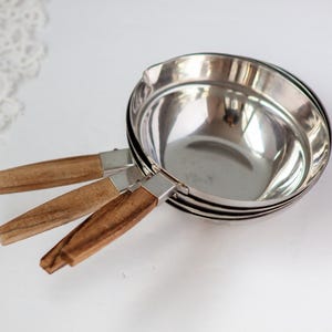 May include: Set of three stainless steel measuring cups with wooden handles. The cups are nested together, showcasing their varying sizes. The handles are a light brown color, and the cups have a shiny, reflective surface.