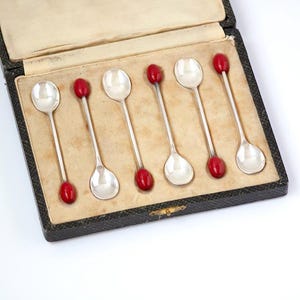 May include: Set of six silver spoons with red accents, presented in a black case with a beige interior. The spoons have a classic design, perfect for serving small portions.