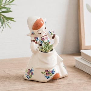 May include: A ceramic figurine of a girl in a Dutch outfit, holding a small succulent plant. The figurine is painted in cream, with floral accents in blue, purple, and orange. The girl's hat and shoes are orange.