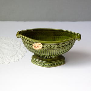 May include: Green ceramic Capri planter with a pedestal base. The planter has a decorative design around the rim and base, with vertical lines on the body. A white lace doily is to the left of the planter.