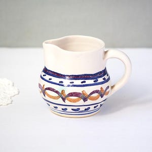 May include: A small, off-white ceramic pitcher with a handle and spout. The pitcher is decorated with blue, purple, and orange geometric patterns around the body. The design includes a band of blue dots and a band of purple and orange interlocking shapes.