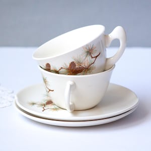 May include: Two cream-colored teacups stacked on saucers. Each cup and saucer features a pine cone and branch design in brown and green. The cups have curved handles.