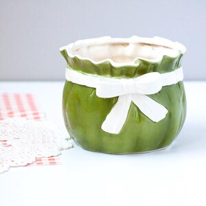 May include: A glossy olive green ceramic planter with a ruffled top edge and a sculpted white ribbon tied around the center. The planter has a rounded, bulbous shape with a smooth, reflective finish and a wide opening at the top for plants.