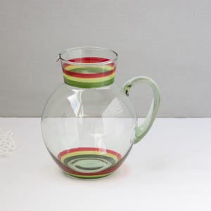 May include: A clear glass pitcher with a round body and a curved handle. The pitcher is adorned with horizontal bands of red, yellow, and green. The matching lid also features the same colorful stripes. The pitcher is suitable for serving beverages.