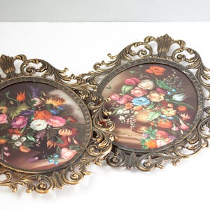 May include: Two vintage floral wall art pieces. Each features a round print of a colorful flower arrangement in a vase. The prints are set in ornate, gold-toned, oval frames with intricate scrollwork.