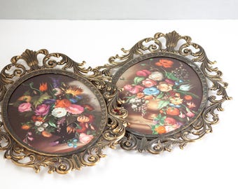 May include: Two vintage floral wall art pieces. Each features a round print of a colorful flower arrangement in a vase. The prints are set in ornate, gold-toned, oval frames with intricate scrollwork.