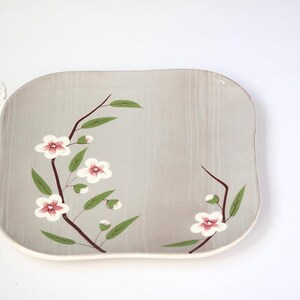 May include: A square, light gray ceramic serving platter with rounded corners. The platter is decorated with a floral design featuring white flowers with pink centers, green leaves, and brown stems. The rim is off-white.
