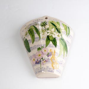 May include: Cream-colored ceramic wall pocket with a floral and musical note design. The pocket features green leaves, white flowers, and purple flowers. A yellow violin is also visible.