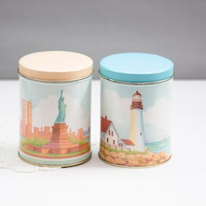 May include: Two round metal storage tins. One tin features the Statue of Liberty and a cityscape, the other a lighthouse scene. Both have a light blue and tan lid, respectively. The tins are cylindrical and have a vintage aesthetic.