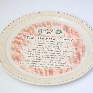 May include: A round ceramic plate with a cream-colored base and a yellow dashed rim. The center features a red cross-hatched background with a cartoon cookie illustration and the text: The Thankful Cookie, I am a thankful cookie, a thankful cookie I be, I'm thankful I can bring a smile to any face I see, I'm thankful I can make people feel good for all the kind things they do, I'm thankful I can cheer anyone up on a day they're feeling blue, I'm thankful to be sweet, even sweeter when I am shared, I'm the perfect way to let someone know just how much you care.
