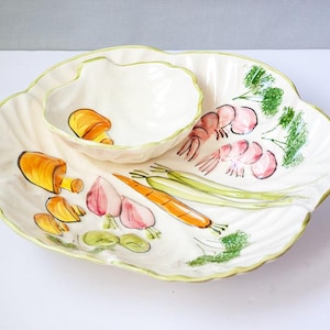 May include: A white ceramic serving platter with a scalloped edge and a small, attached bowl. The platter is decorated with hand-painted illustrations of vegetables and shrimp, with a green trim.