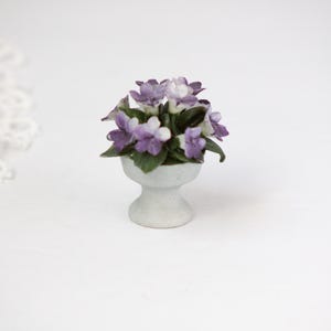 May include: A miniature white pedestal planter filled with purple and white flowers and green leaves. The small ceramic pot is centered on a white surface. The flowers are in full bloom, creating a delicate floral arrangement.