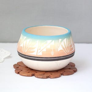 May include: A rounded ceramic bowl featuring a gradient design that transitions from a light blue rim to a soft peach midsection. The exterior is decorated with white geometric leaf patterns and a solid black horizontal band near the base.