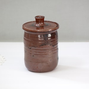 May include: A handmade, brown ceramic jar with a lid. The jar has a cylindrical shape with a slightly textured surface and a small handle on the lid. The jar is likely used for storage and has a rustic, earthy aesthetic.