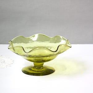 May include: A light green, ruffled-edge glass bowl on a pedestal base. The bowl has a textured surface and a scalloped rim. The glass has a slight yellow tint, and the bowl is likely vintage or antique.