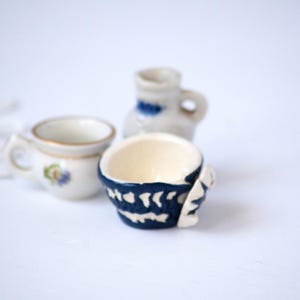 May include: Three small ceramic pieces: a cup with a handle and floral design, a small pitcher with blue accents, and a blue and white patterned bowl. The items are on a white surface.