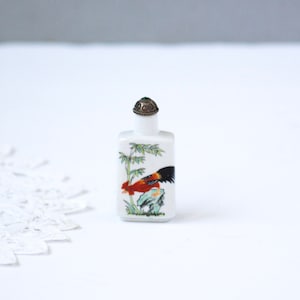 May include: A small, rectangular white ceramic bottle with a detailed painting of a rooster and bamboo. The rooster is red, black, and orange. The bottle has a decorative silver-colored stopper with a green gem.