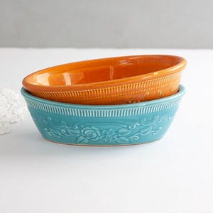 May include: Two oval ceramic bowls stacked together. The top bowl is a vibrant orange, while the bottom bowl is a bright turquoise. Both bowls feature a decorative embossed floral pattern around the sides and a vertical ribbed rim detail.