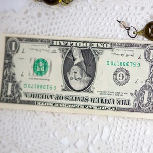 K&ouml;nnte beinhalten: Ein Ein-Dollar-Schein mit dem Portr&auml;t von George Washington, der Seriennummer C51388170D und dem Text "THE UNITED STATES OF AMERICA". Der Schein liegt auf einer wei&szlig;en, spitzenartigen Unterlage, mit einer dekorativen Glasperle in der N&auml;he.