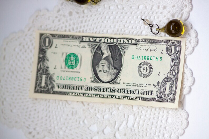K&ouml;nnte beinhalten: Ein Ein-Dollar-Schein mit dem Portr&auml;t von George Washington, der Seriennummer C51388170D und dem Text "THE UNITED STATES OF AMERICA". Der Schein liegt auf einer wei&szlig;en, spitzenartigen Unterlage, mit einer dekorativen Glasperle in der N&auml;he.