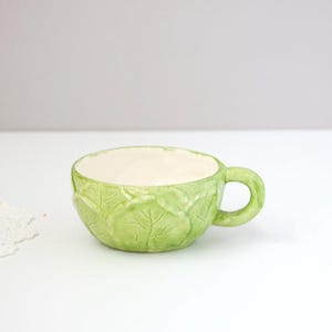 May include: A ceramic cup with a light green exterior featuring a molded cabbage leaf texture and a smooth, off-white interior. The cup has a rounded handle on the right side and a wide, shallow bowl shape, showcasing detailed leaf veins.
