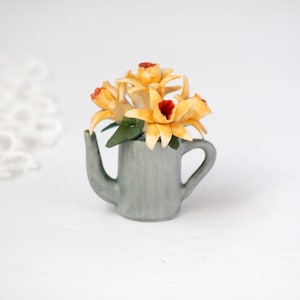 May include: A miniature gray watering can filled with yellow and red flowers. The flowers have green leaves and are arranged in a small, decorative container. The background is white.