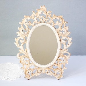 May include: An off-white, ornate oval mirror with a decorative, scrolling frame. The mirror is freestanding, with a distressed finish, and sits on a white surface. A white lace doily is in the foreground. The mirror is a decorative home accent.