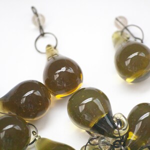 K&ouml;nnte beinhalten: Nahaufnahme von mehreren olivgr&uuml;nen, tropfenf&ouml;rmigen Glasperlen mit kleinen Metallschlaufen und klaren Perlen. Die Perlen sind auf einer wei&szlig;en Oberfl&auml;che angeordnet. Die Glasperlen haben eine gl&auml;nzende Oberfl&auml;che.