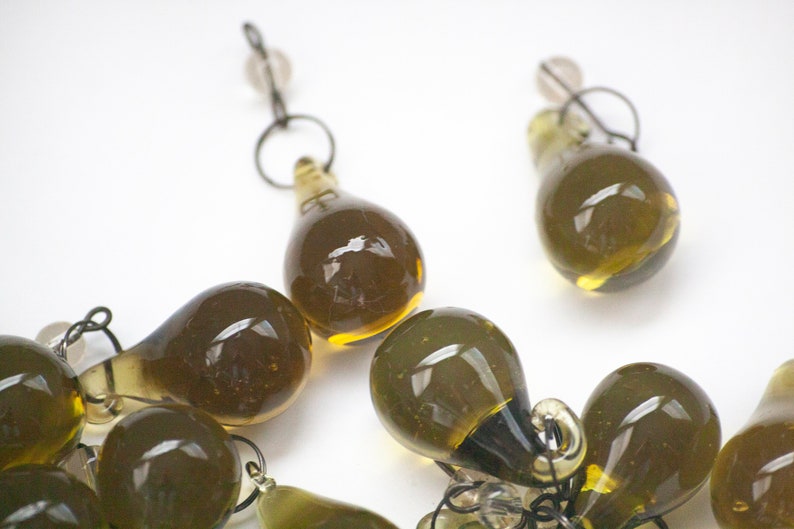 K&ouml;nnte beinhalten: Nahaufnahme von mehreren olivgr&uuml;nen, tropfenf&ouml;rmigen Glasperlen mit kleinen Metallschlaufen und klaren Perlen. Die Perlen sind auf einer wei&szlig;en Oberfl&auml;che angeordnet. Die Glasperlen haben eine gl&auml;nzende Oberfl&auml;che.