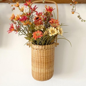 May include: A woven wicker basket filled with colorful artificial flowers in shades of orange, red, and yellow. The basket has a handle and is hanging on a wooden bar. Decorative beaded garland is above the basket.
