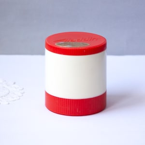 May include: A cylindrical container with a red top and base, and a white central section. The top has the word "Aladdin" printed on it. The container is on a white surface.