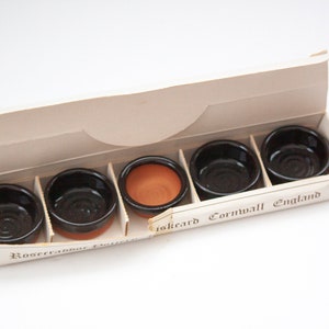 May include: A set of five small, round, black ceramic bowls with a terracotta-colored interior, presented in a cardboard box. The box is labeled with text that reads "Liskeard Cornwall England".
