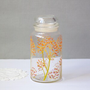 May include: A clear glass jar with a lid, decorated with a yellow and orange floral design. The jar is cylindrical and sits on a white surface. A white doily is in the lower left corner.