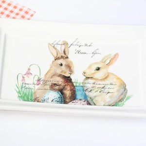 May include: Rectangular white ceramic serving dish with handles. The dish features a watercolor illustration of two rabbits, Easter eggs, and floral accents. Decorative script is printed across the top.