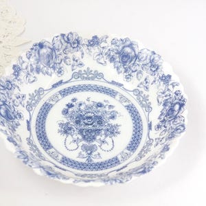 May include: White ceramic bowl with a blue floral design. The bowl has a scalloped edge and a detailed pattern of flowers and leaves. The center of the bowl features a bouquet of flowers. A white lace doily is in the background.